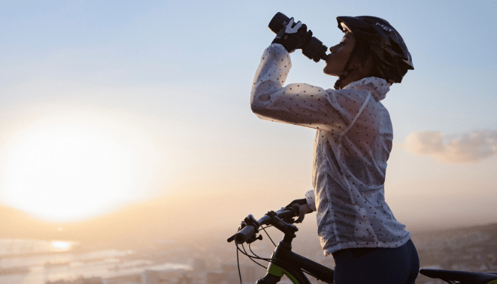 Woman drinking from bottle while holding bike at sunset