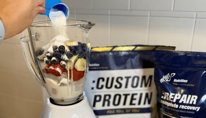 Pouring protein into a blender with ice, strawberrys, blueberrys, banana's ice, and almond milk